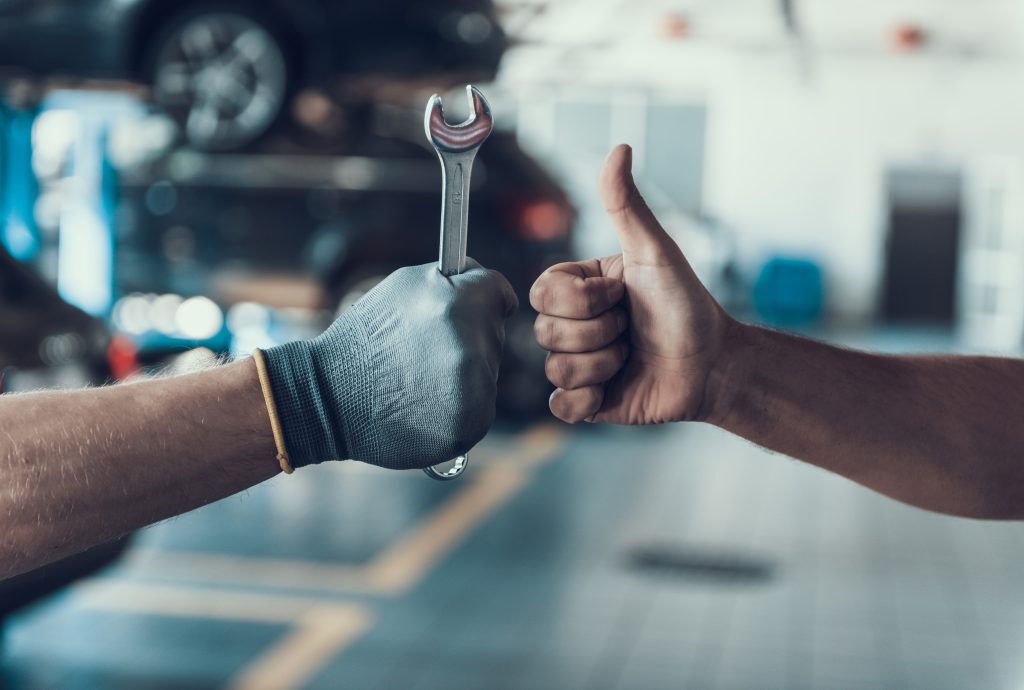 A mechanic handing over a wrench to a colleague with a thumbs up gesture, symbolizing teamwork and quality service in an auto repair shop.