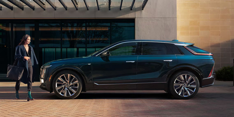 Woman walking toward a dark blue Cadillac LYRIQ SUV parked outside a modern building.