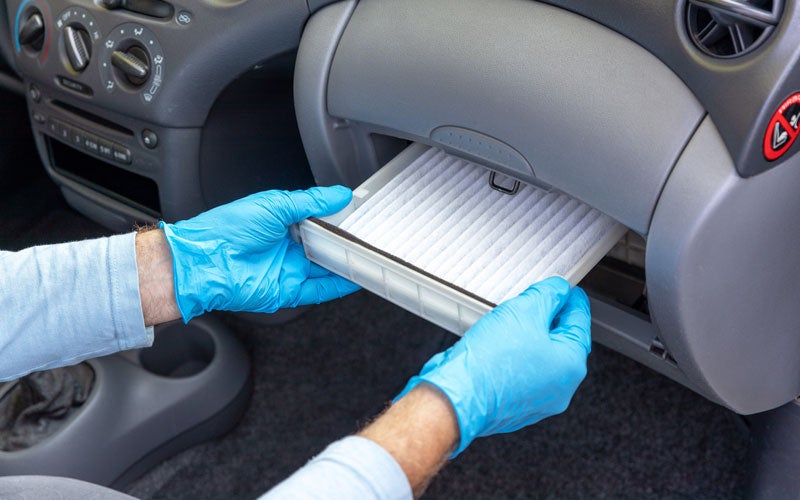 Person in blue gloves changing a car's cabin air filter, positioned behind the glove compartment.