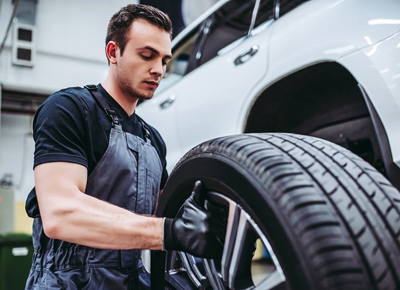 A male mechanic in gray overalls and black gloves holds a car tire. A white SUV is in the background.