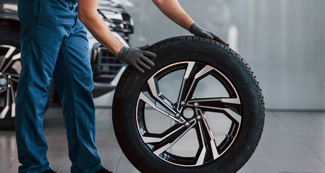 A mechanic, wearing blue coveralls and dark gloves, rolls a new car tire with a shiny silver and black rim.
