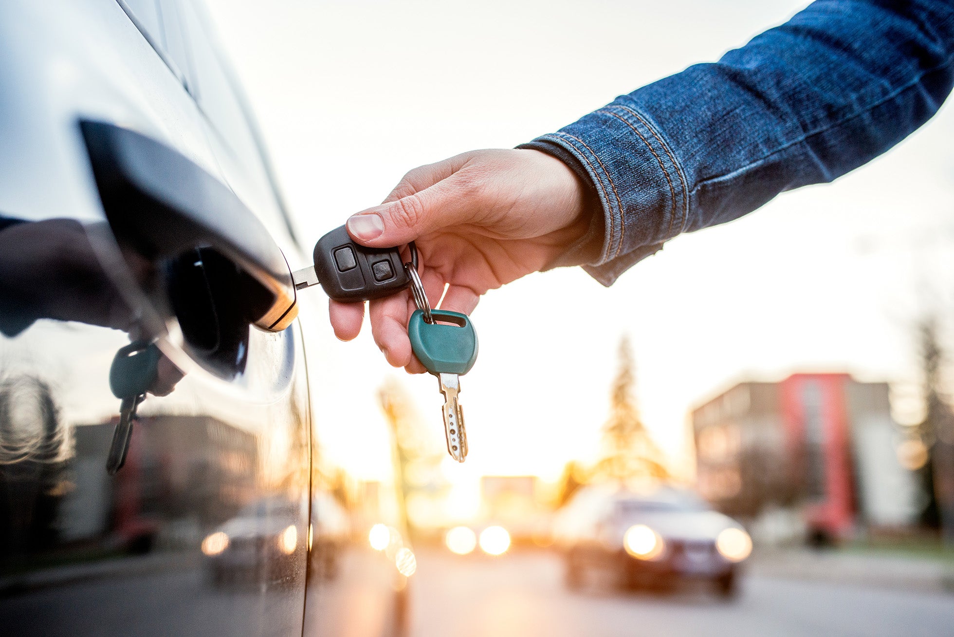 A person in a denim jacket putting a car key into the door lock of a dark car, with sunset glare.