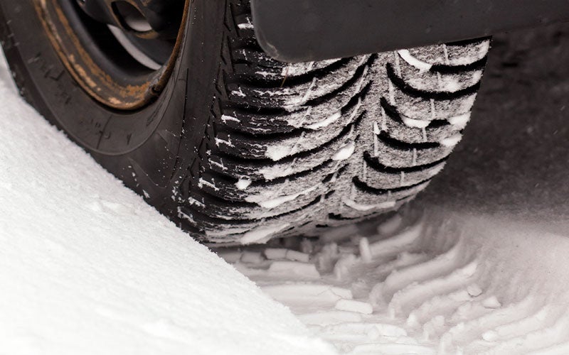 A car tire, with thick tread covered in snow, spinning in deep snow and leaving a track.